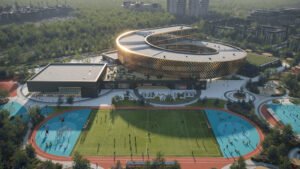 Low-angle aerial architectural render focusing on the school’s outdoor sports facilities, including a large green football pitch with a running track, colorful blue play courts, and an adjacent rectangular sports block building. The main spiral school building is visible in the background.