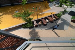 A man sits on a wooden block bench while two young girls play on nearby boulders and benches in a lushly planted plaza. The space is surrounded by native Plumeria trees and dense green shrubbery, illustrating how the campus design serves as a comfortable, shared hub for students, faculty, and families.