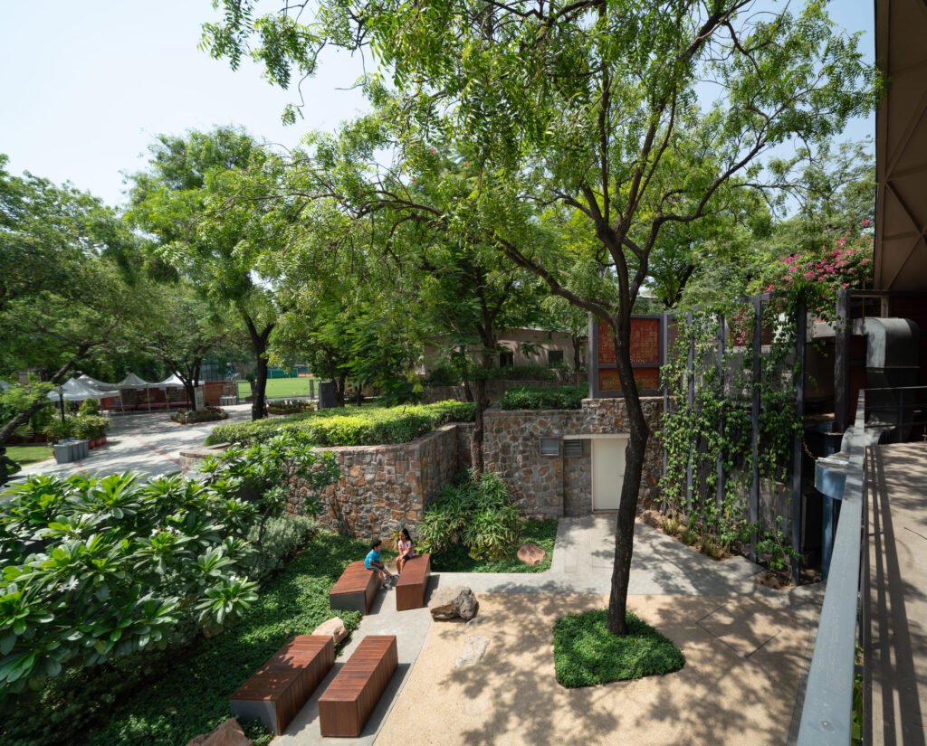 Looking out from an elevated walkway over the school courtyard. A man leans on the metal railing, looking down at the sandstone paths, native dry scrub plantings, and a fabric sunshade. In the background, modern school buildings with brick accents rise amongst mature green trees.