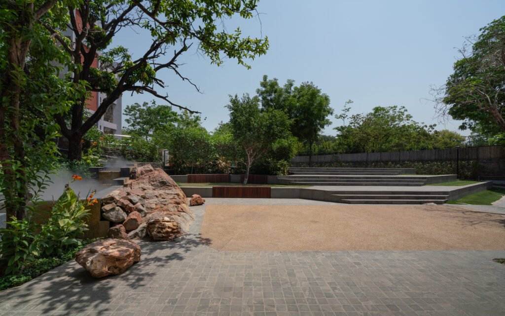  A wide view of a spacious, manicured green grass lawn punctuated by large, rugged boulders. A curved slate pathway lines the left side of the lawn, while yellow basketball hoops and courts are visible in the background. The open softscape design maximizes ground permeability for rainwater harvesting.