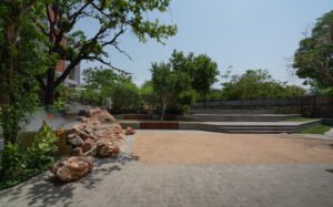 A wide view of a spacious, manicured green grass lawn punctuated by large, rugged boulders. A curved slate pathway lines the left side of the lawn, while yellow basketball hoops and courts are visible in the background. The open softscape design maximizes ground permeability for rainwater harvesting.