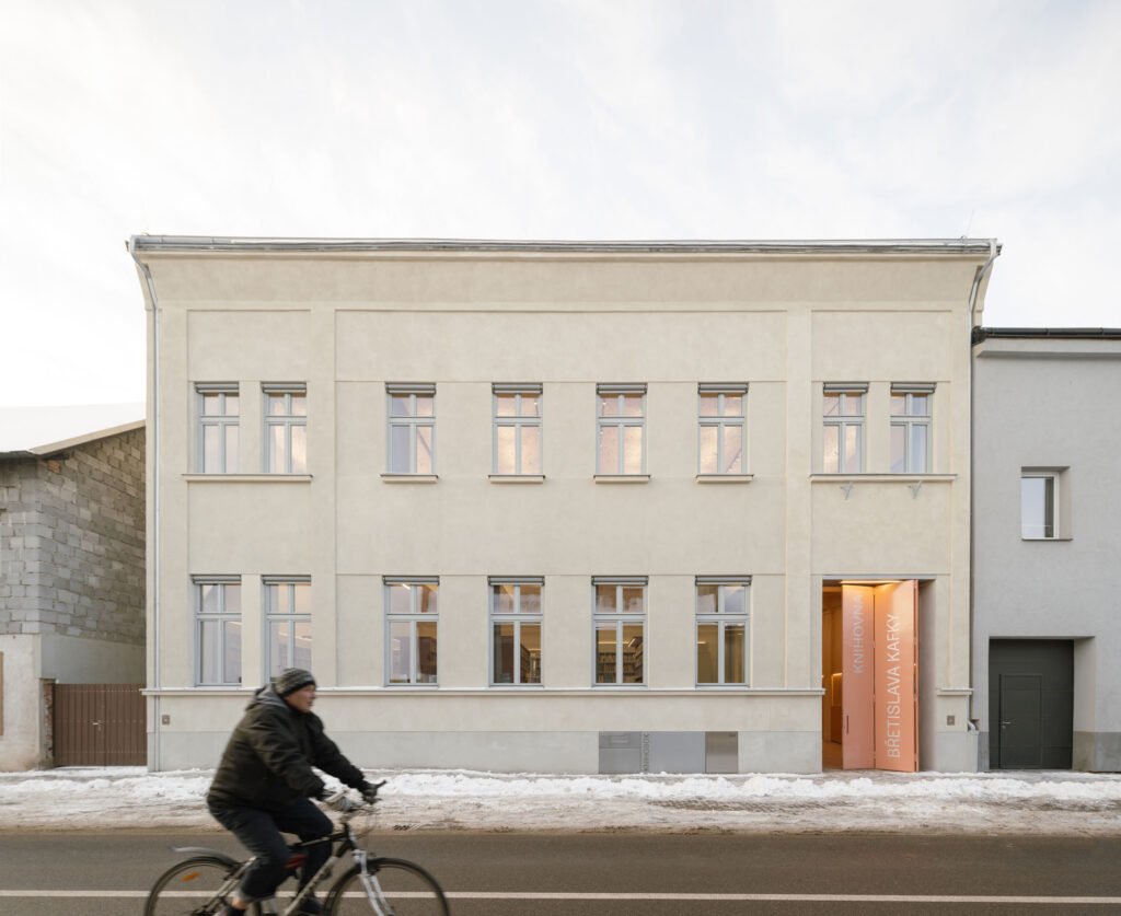 Exterior street-side view of the historic Břetislav Kafka Library in winter, showing its clean beige plastered facade with grey window frames and a cyclist passing by on Sokolská street.