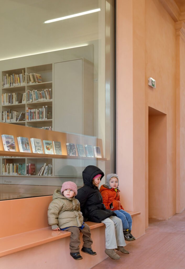 Wide photo of the children's reading room under a wavy white fabric canopy custom-printed with drawings, showcasing pink upholstered mobile benches and small children reading books.