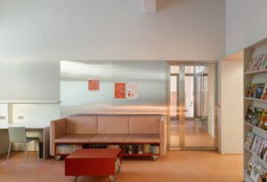 View of the playful kids' library room with varied low modular red and pink furniture on wheels, a tiered wooden seating platform, and a printed textile canopy overhead.