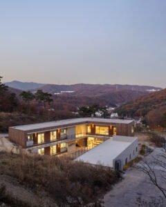 High-angle twilight photograph of the school's L-shaped concrete and wood building, nestled into a steep slope. In the background, the rolling, forested hills of the Gogi-dong valley stretch out under a clear dusk sky.