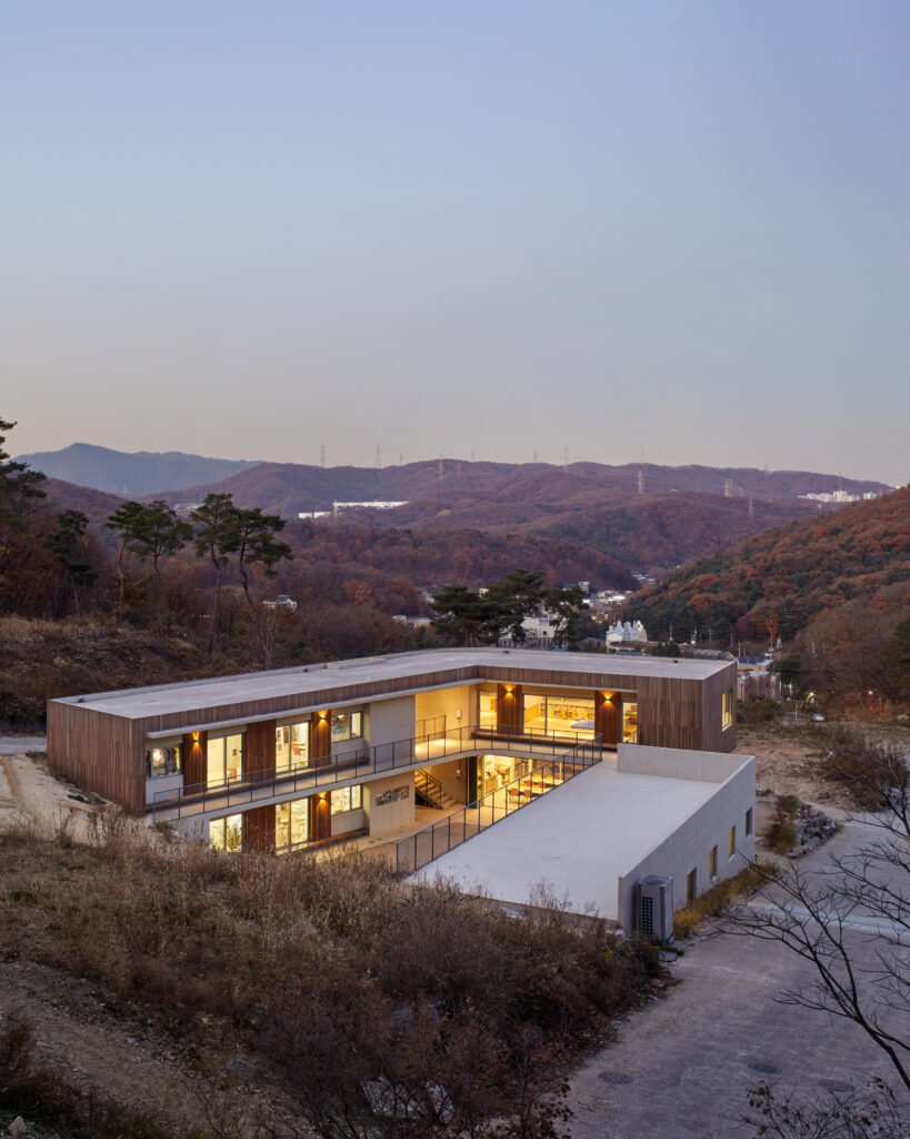 High-angle twilight photograph of the school's L-shaped concrete and wood building, nestled into a steep slope. In the background, the rolling, forested hills of the Gogi-dong valley stretch out under a clear dusk sky.