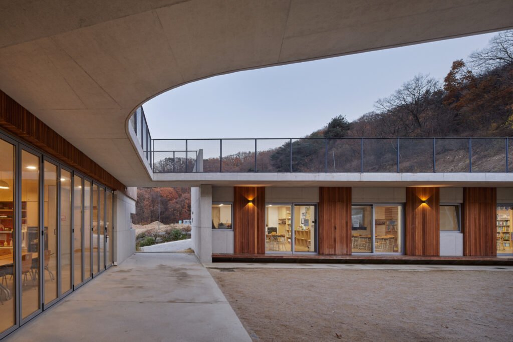Twilight view of the courtyard looking underneath a cantilevered, raw concrete second-floor corridor. The concrete slab forms a protective canopy over the traditional wooden toenmaru (narrow porch) lining the ground-floor classrooms.