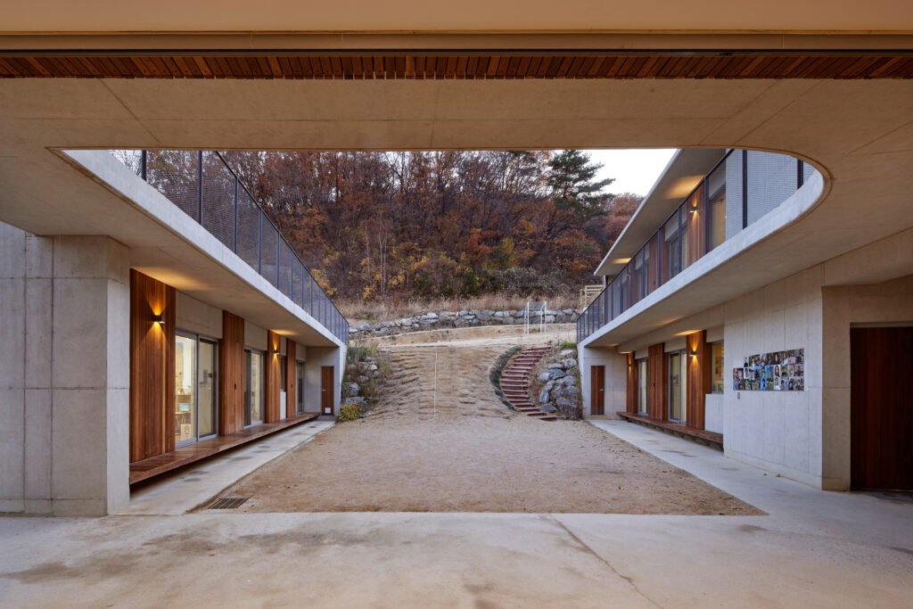 Twilight shot from within the central courtyard looking upward. A wide, unpaved sloped path and curved stone-and-brick steps rise steeply between the two building blocks, leading directly into the Gwang-gyo mountain forest.