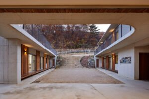 Twilight shot from within the central courtyard looking upward. A wide, unpaved sloped path and curved stone-and-brick steps rise steeply between the two building blocks, leading directly into the Gwang-gyo mountain forest.