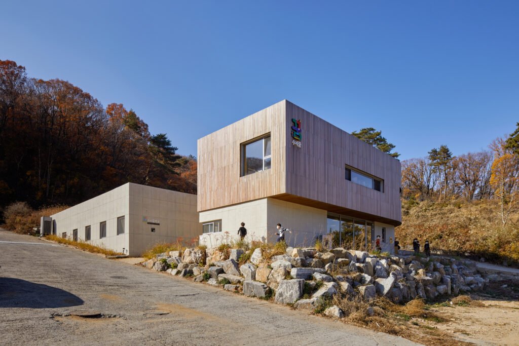 Daytime photograph of the school’s front entrance under a clear blue sky, highlighting a concrete lower level and a wooden-clad upper volume with the school logo. Children play near the stone embankment on the right.