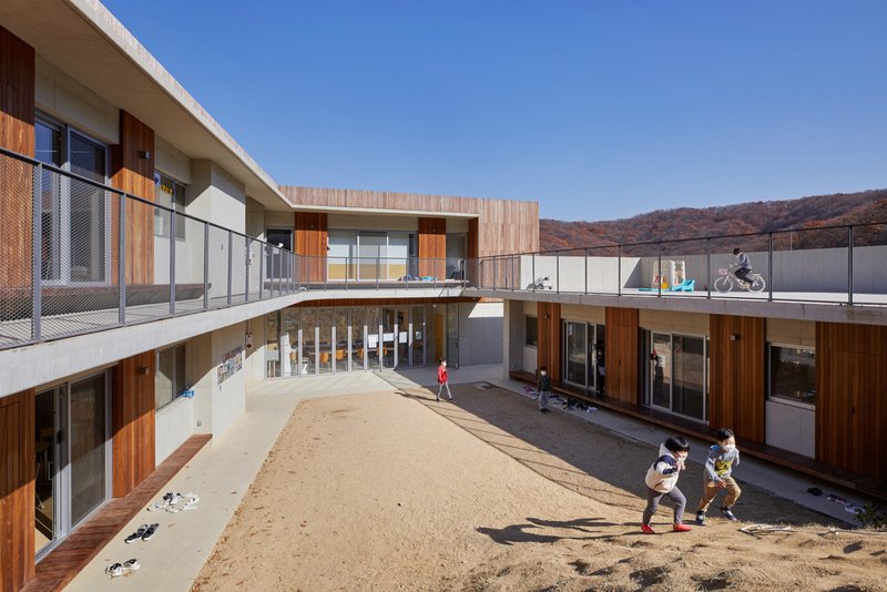 Dynamic, high-angle daytime view of children running and playing across the sandy courtyard, the sloped hillside playground, and the wide concrete roof deck, showing how the building wraps around the active central space.