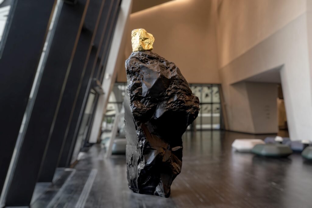 Sculptural artwork of a rugged, charcoal-black stone pillar topped with a gleaming chunk of polished gold.