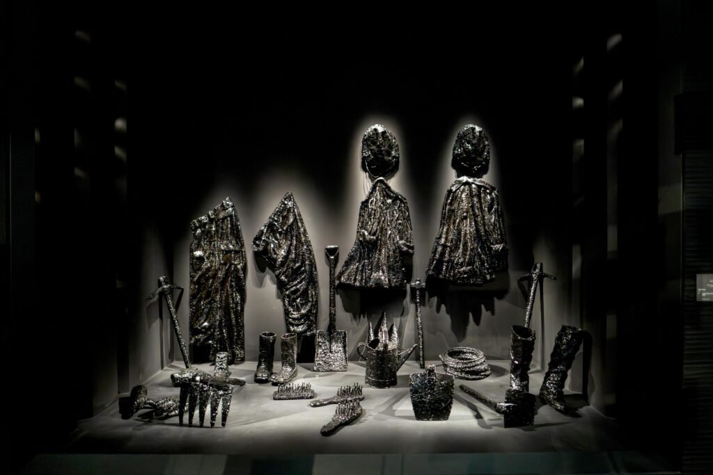 Exhibition showcase of everyday clothes, tools, and shovels entirely coated in a thick, shiny black oil-like crust.