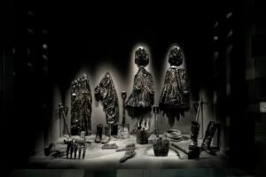 Exhibition showcase of everyday clothes, tools, and shovels entirely coated in a thick, shiny black oil-like crust.