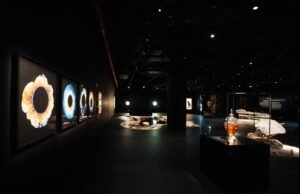 Dark gallery displaying backlit, circular eye iris artwork and a glowing glass case with a jar of amber liquid.