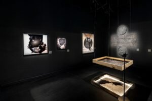 Dark room showing photographs of oil-smeared faces and a suspended wooden tray displaying tiny vertical pins.