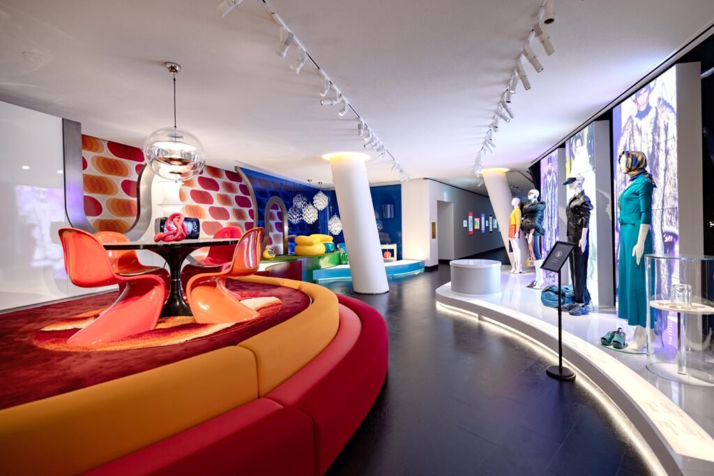 1970s interior with bright orange plastic chairs, circular patterns, and colourful mannequins.