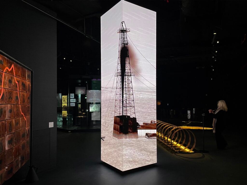 A tall, three-sided LED pillar displaying video of a black plume gushing from an industrial oil derrick.