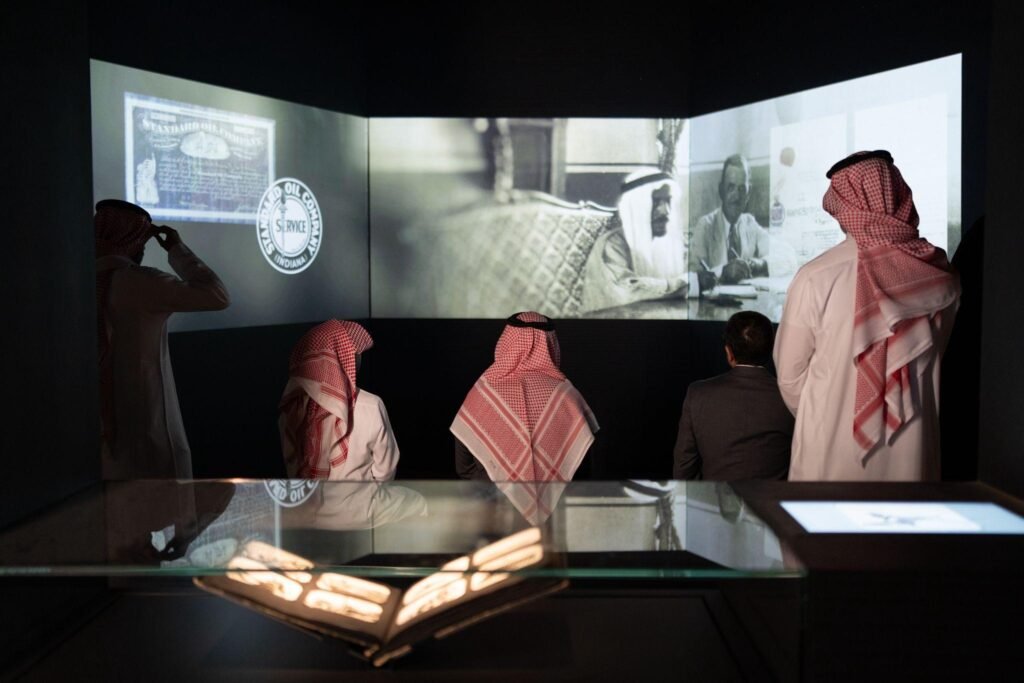 Visitors watching a historic black-and-white documentary projection across three massive screens in a dark room.