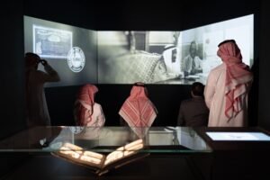 Visitors watching a historic black-and-white documentary projection across three massive screens in a dark room.