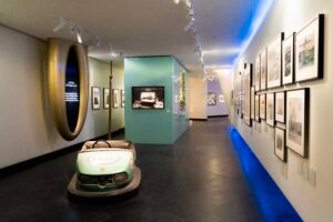 A retro green Esso brand bumper car in a museum gallery with vertical historical photographic panels on walls.