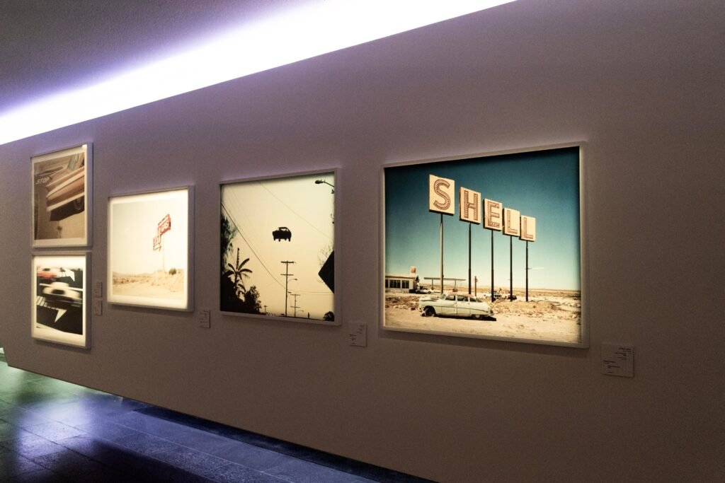 Backlit gallery frames displaying photographs of highway scenes and a classic, brightly lit Shell gas station sign.