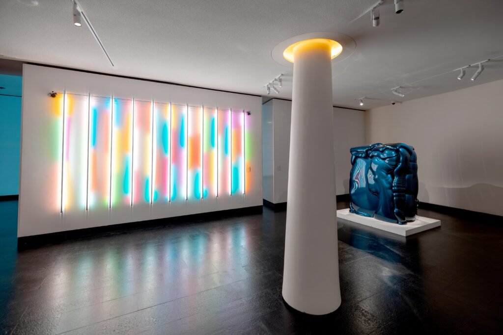 White gallery room featuring pastel vertical neon tube lights, a white column, and a squished metallic blue car sculpture.