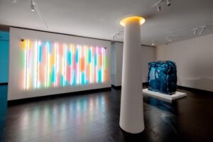 White gallery room featuring pastel vertical neon tube lights, a white column, and a squished metallic blue car sculpture.