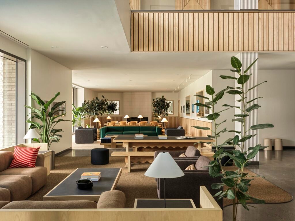 Open-plan lobby and lounge on the ground floor of the Head House, showing fluted columns, natural wood wall panels, a long study table with chairs in the background, potted trees, and modern armchairs on a brown carpet.