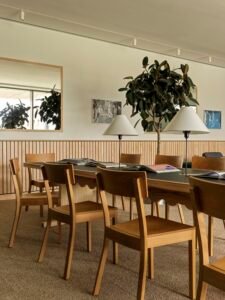 An angled shot of a custom long wooden study table paired with simple wooden chairs and topped with minimalist white-coned table lamps in front of a wood-slat wainscot wall.