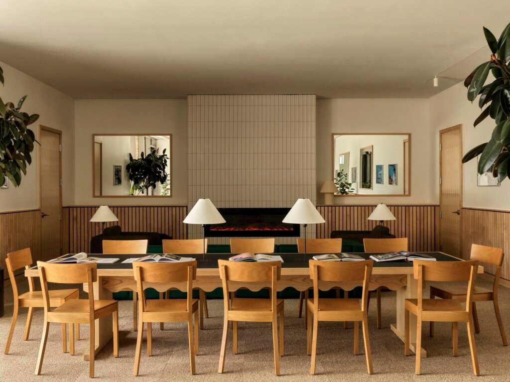 Front view of a long wooden study table with built-in desk lamps and wooden chairs, placed in front of a minimalist fireplace with vertical light-grey tiling and flanked by horizontal mirrors.