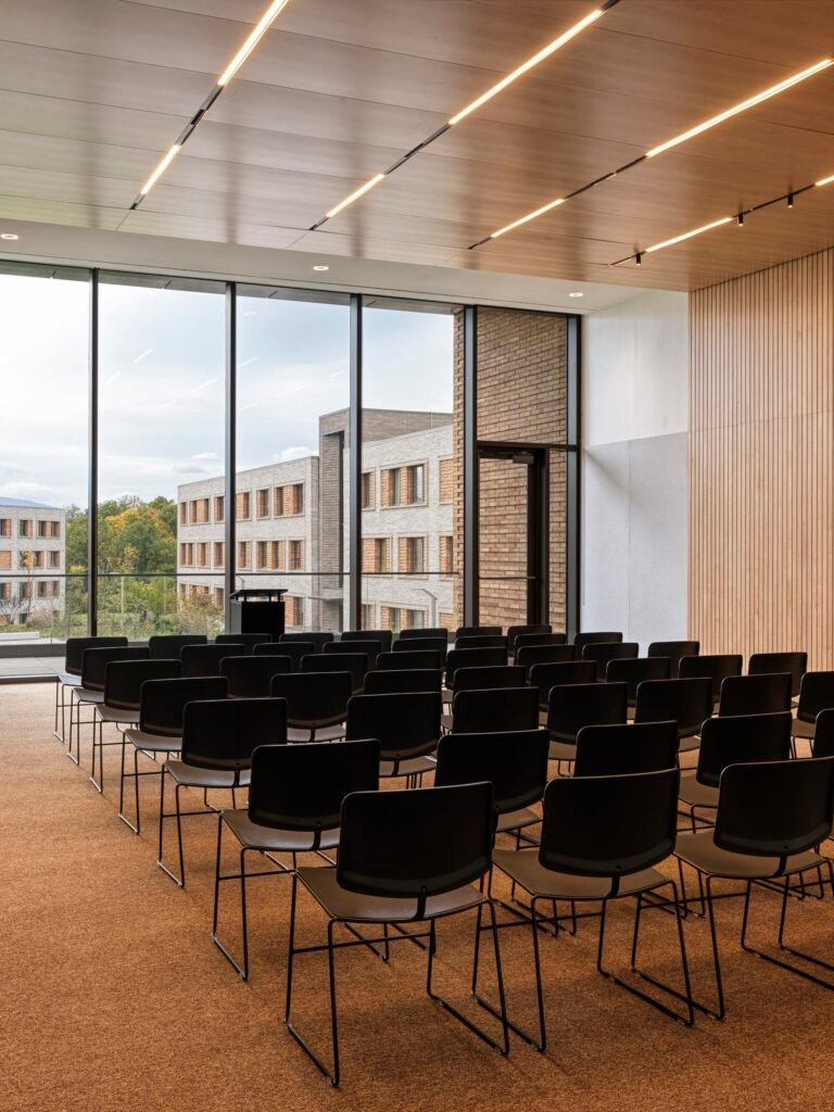 A contemporary lecture or study room containing rows of black chairs facing a speaker's podium. A large glass wall overlooks the exterior brick dormitory buildings.