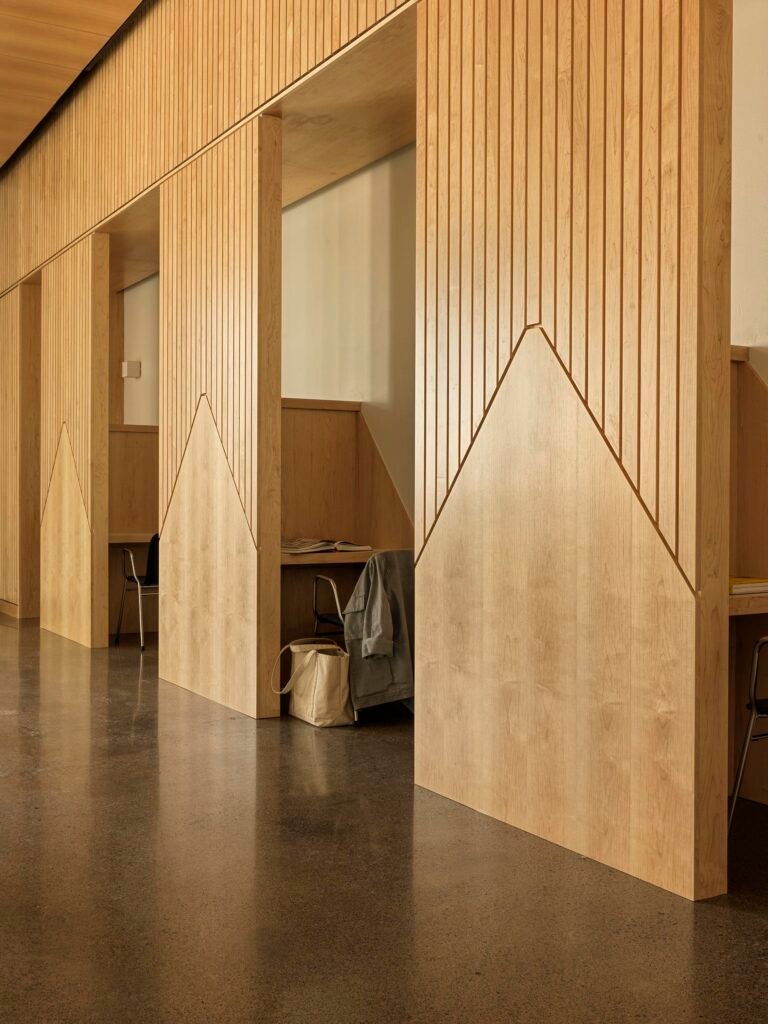 A row of custom wooden study carrels divided by fluted wood screens with geometric mountain-shaped cutouts. A canvas bag hangs off the side of one partition.