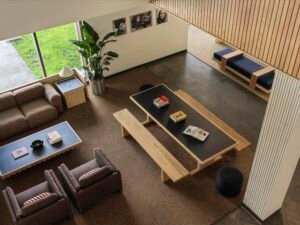 Inside the Head House lounge looking through large floor-to-ceiling windows toward the grassy exterior green and brick residence halls. Features a green modular sofa and a blue upholstered bench.