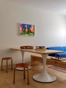 Interior of a student apartment showing a dining table with white trumpet pedestals, a solid wooden bench, and round stools. A blue wool sofa and a framed piece of colourful art are in the background.