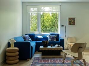 A student apartment common living space with a blue modular corner sofa, a black wavy-edged coffee table, a stacked wooden disc side table, and wooden chairs over a patterned red rug.