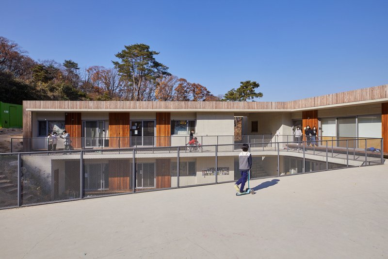 A student rides a blue scooter on the wide, concrete second-floor deck under a bright blue sky. Open classroom glass doors line the wood-clad structure, creating an open transition between indoor learning and outdoor recreation.