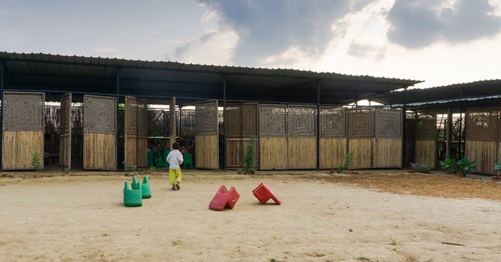 Wide exterior view of the school building featuring a corrugated metal roof and modular charpai woven wall panels facing a wide dirt courtyard with scattered colorful chairs and a child walking toward open doors.