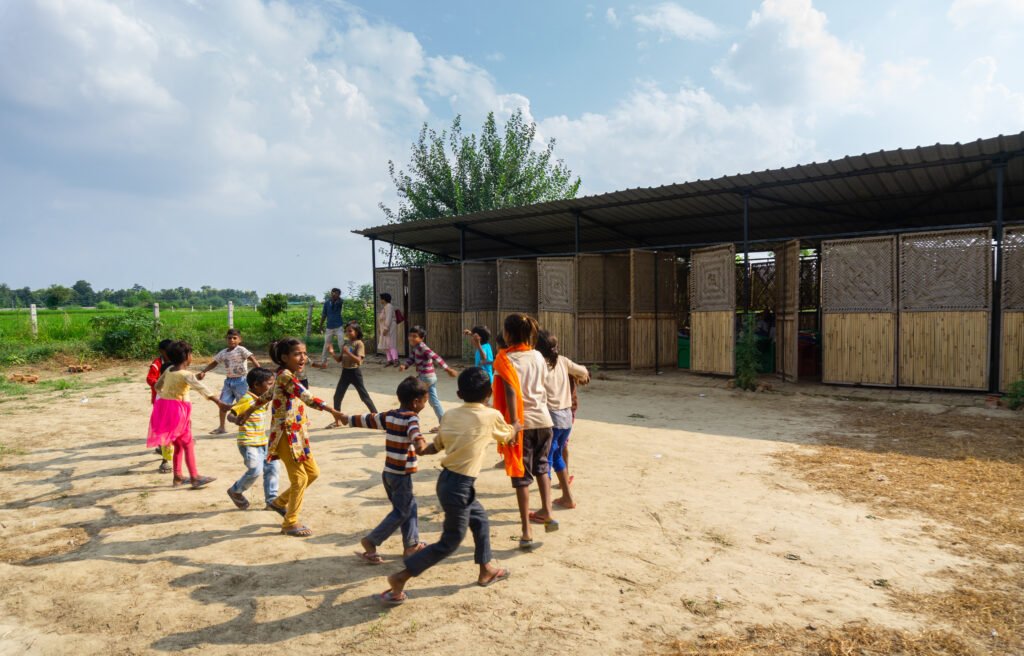 A group of school children holding hands in a circle and playing outdoors in a dirt courtyard in front of the long, modular school structure.