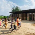 A group of school children holding hands in a circle and playing outdoors in a dirt courtyard in front of the long, modular school structure.