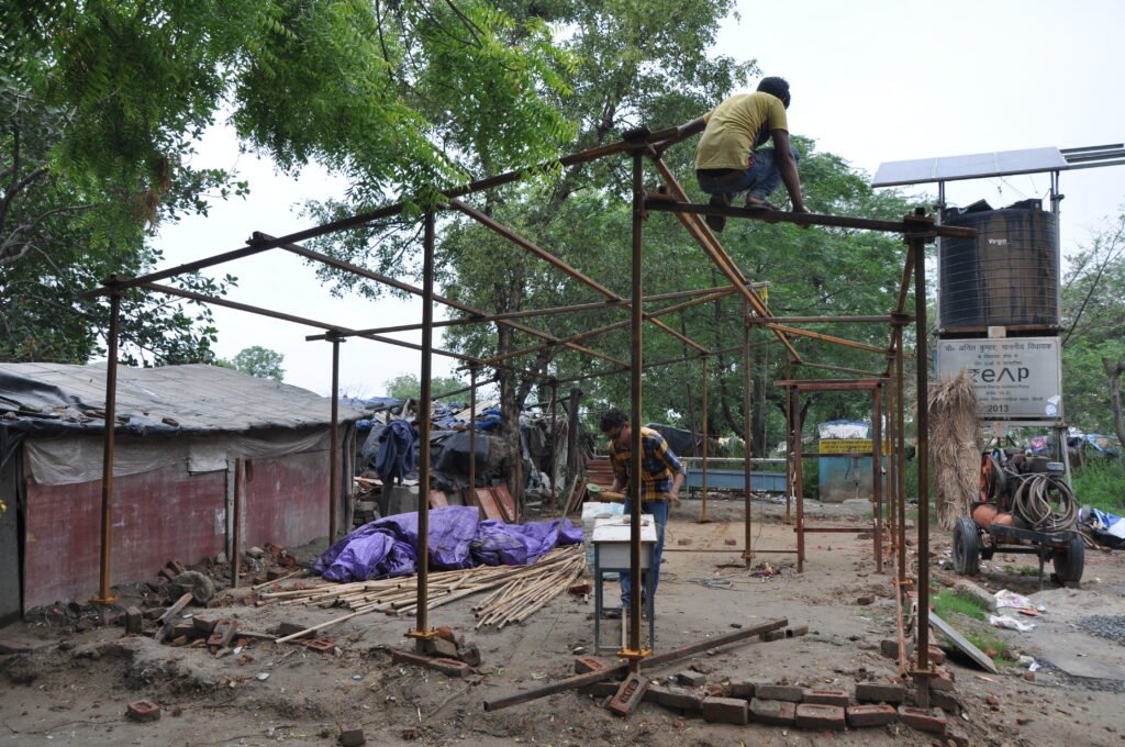 Construction workers assembling a skeletal steel frame structure outdoors, with one worker perching on top of high beams and another working at ground level near stacked bamboo and temporary shelters.