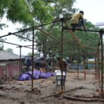 Construction workers assembling a skeletal steel frame structure outdoors, with one worker perching on top of high beams and another working at ground level near stacked bamboo and temporary shelters.
