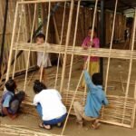 Children working together under a covered frame to thread thin bamboo slats horizontally into standing wooden frame panels.