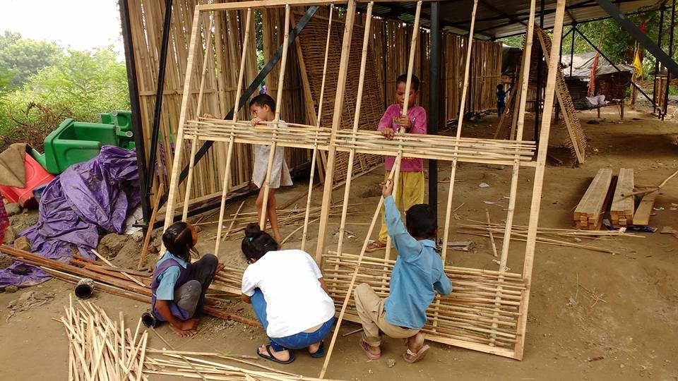 Children working together under a covered frame to thread thin bamboo slats horizontally into standing wooden frame panels.