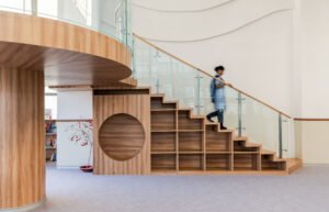 Wooden staircase integrated with open shelf storage boxes and a circular play cutout beneath a glass-railed loft at Vedanya School.