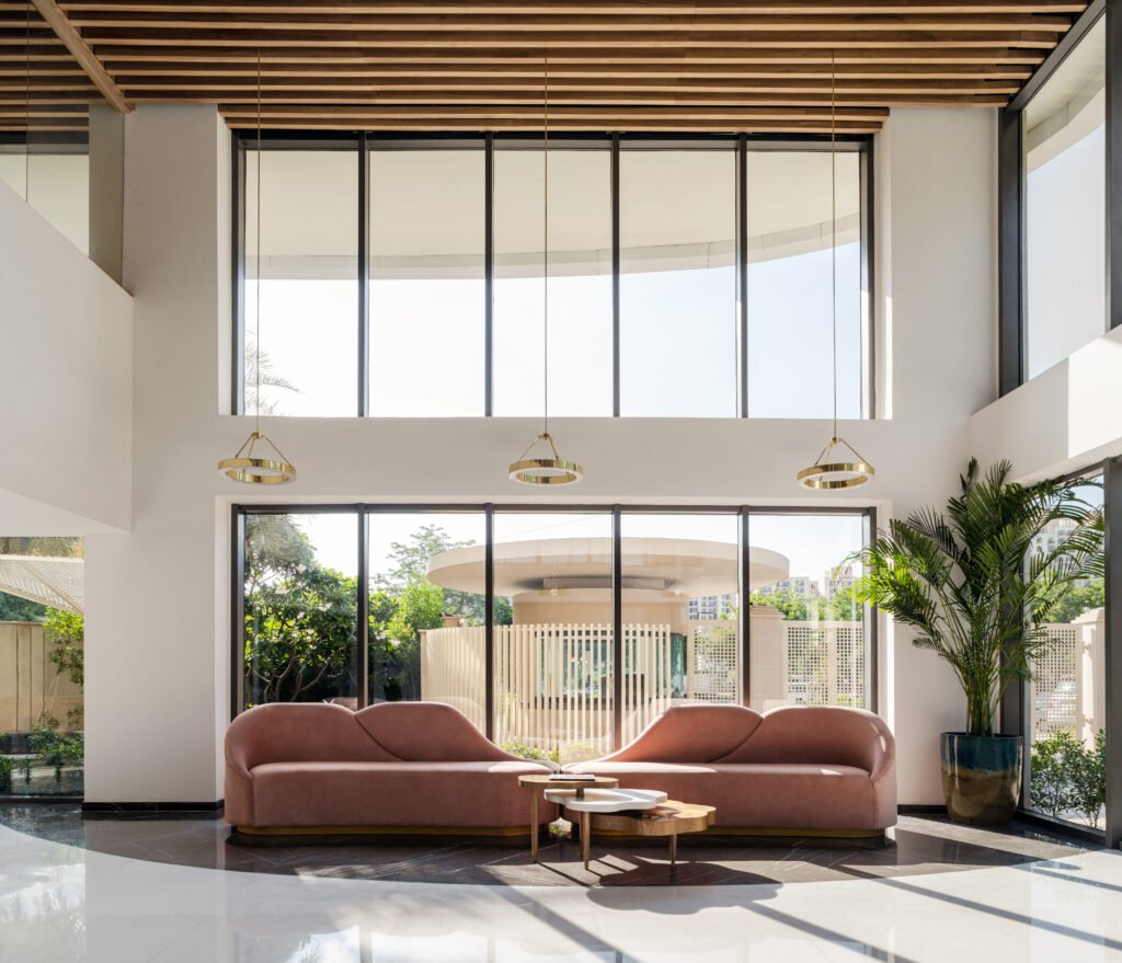 Spacious double-height reception seating area at Vedanya School Gurugram with organic curved pink sofas, gold ring pendant light fixtures, and floor-to-ceiling glass windows.
