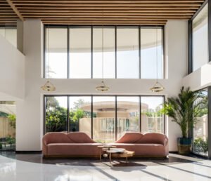 Spacious double-height reception seating area at Vedanya School Gurugram with organic curved pink sofas, gold ring pendant light fixtures, and floor-to-ceiling glass windows.