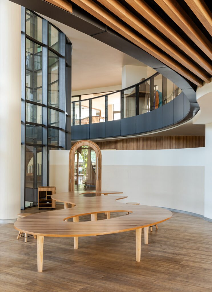 Custom kidney-shaped wooden activity table in a wide curved hallway with slatted timber ceiling soffit and glass atrium walls at Vedanya School.