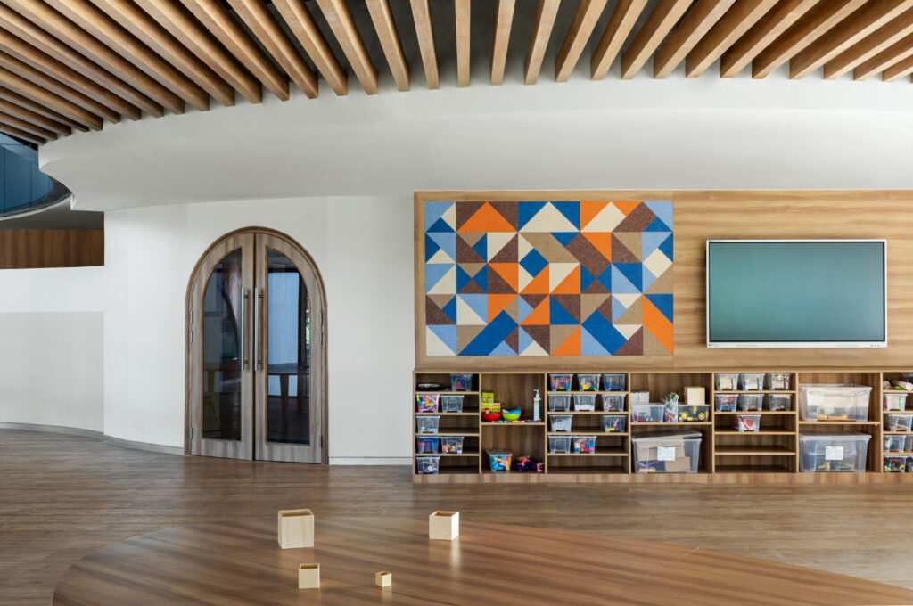 Primary classroom interior at Vedanya School showing a colorful geometric acoustic felt wall panel, interactive digital screen, organized low storage bins, and arched timber door.
