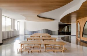 First-floor dining area at Vedanya School featuring light wooden dining tables and benches, blue-and-white striped wall trim, and fluid organic ceiling wave paneling.